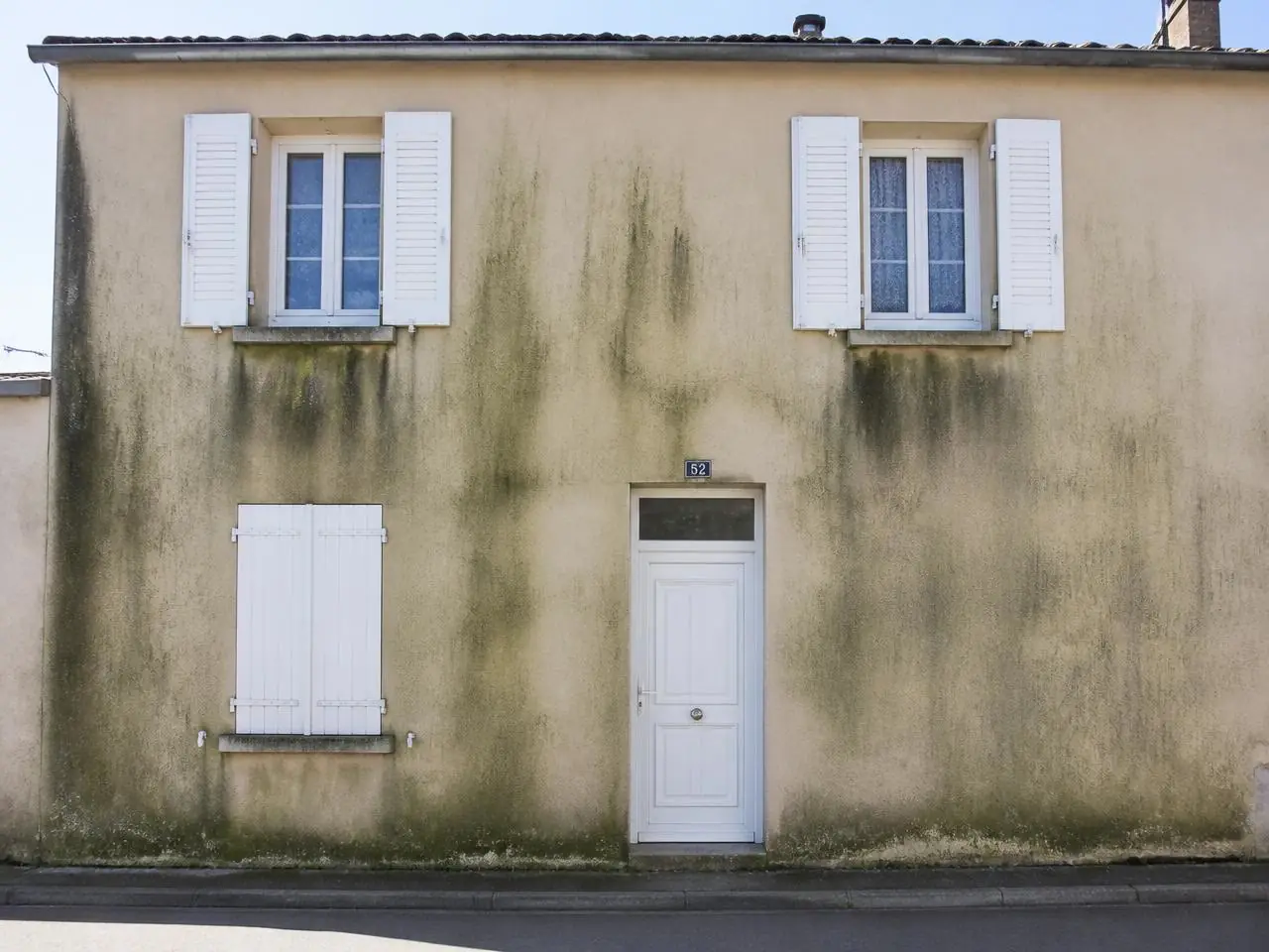 Façade sur rue à l’enduit beige couverte de longues coulures vertes et noires, volets blancs.