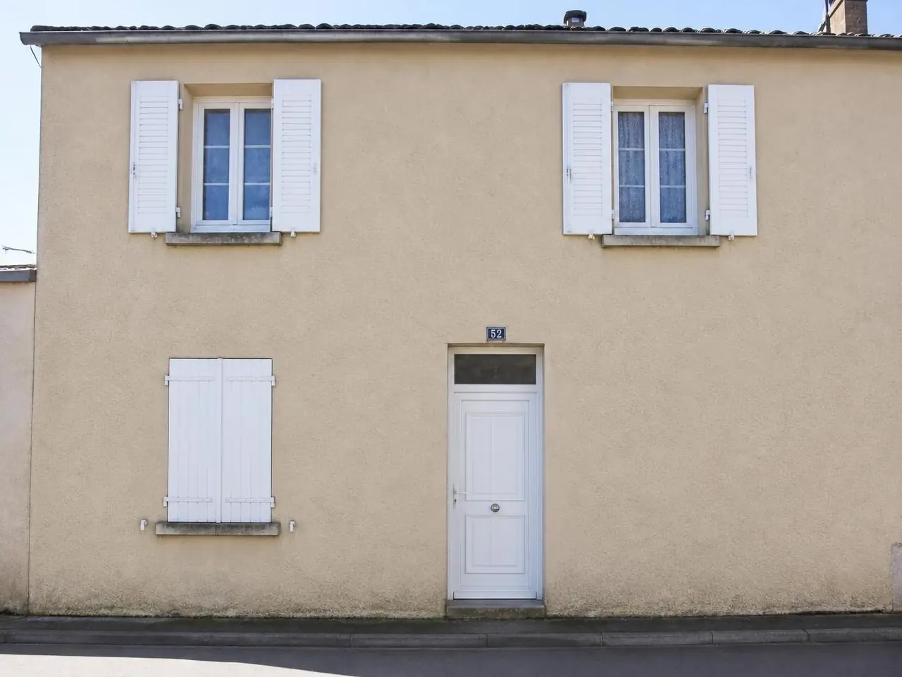 La même façade sur rue, enduit beige net et uniforme sans aucune coulure, mêmes volets blancs.