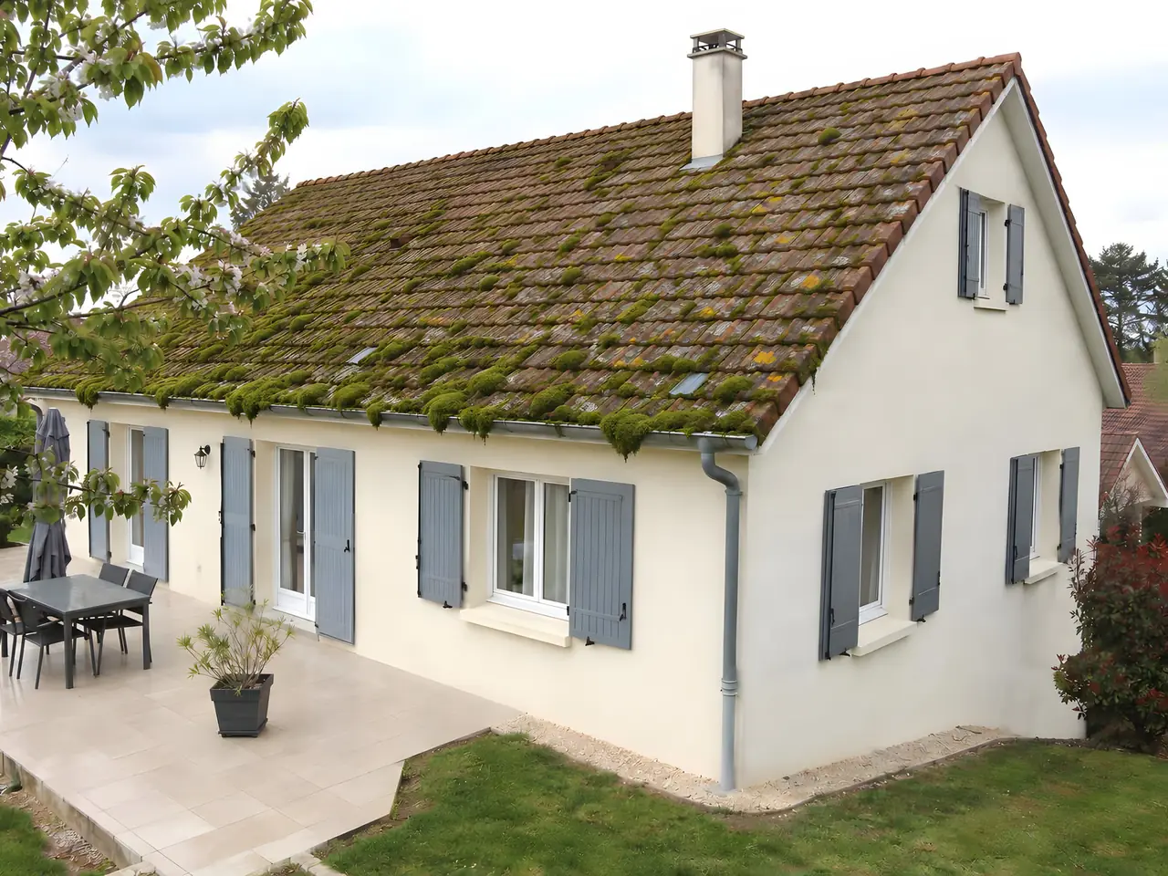 Pavillon au crépi clair et aux volets gris, toit de tuiles couvert de mousse verte jusqu’à la gouttière.